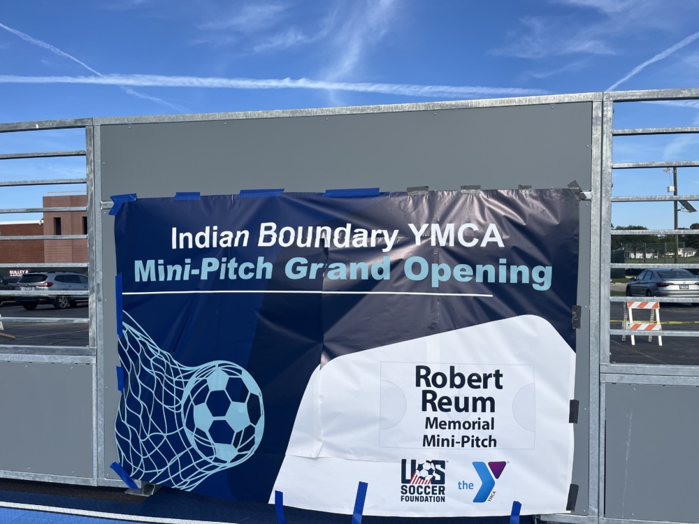 a sky blue, navy blue, and white banner is taped on the face of the goal. It reads "Indian Boundary YMCA Mini-Pitch Grand Opening" and includes the U.S. Soccer Foundation and YMCA logos. It also reads "Robert Reum Memorial Mini-Pitch" in the bottom right corner.